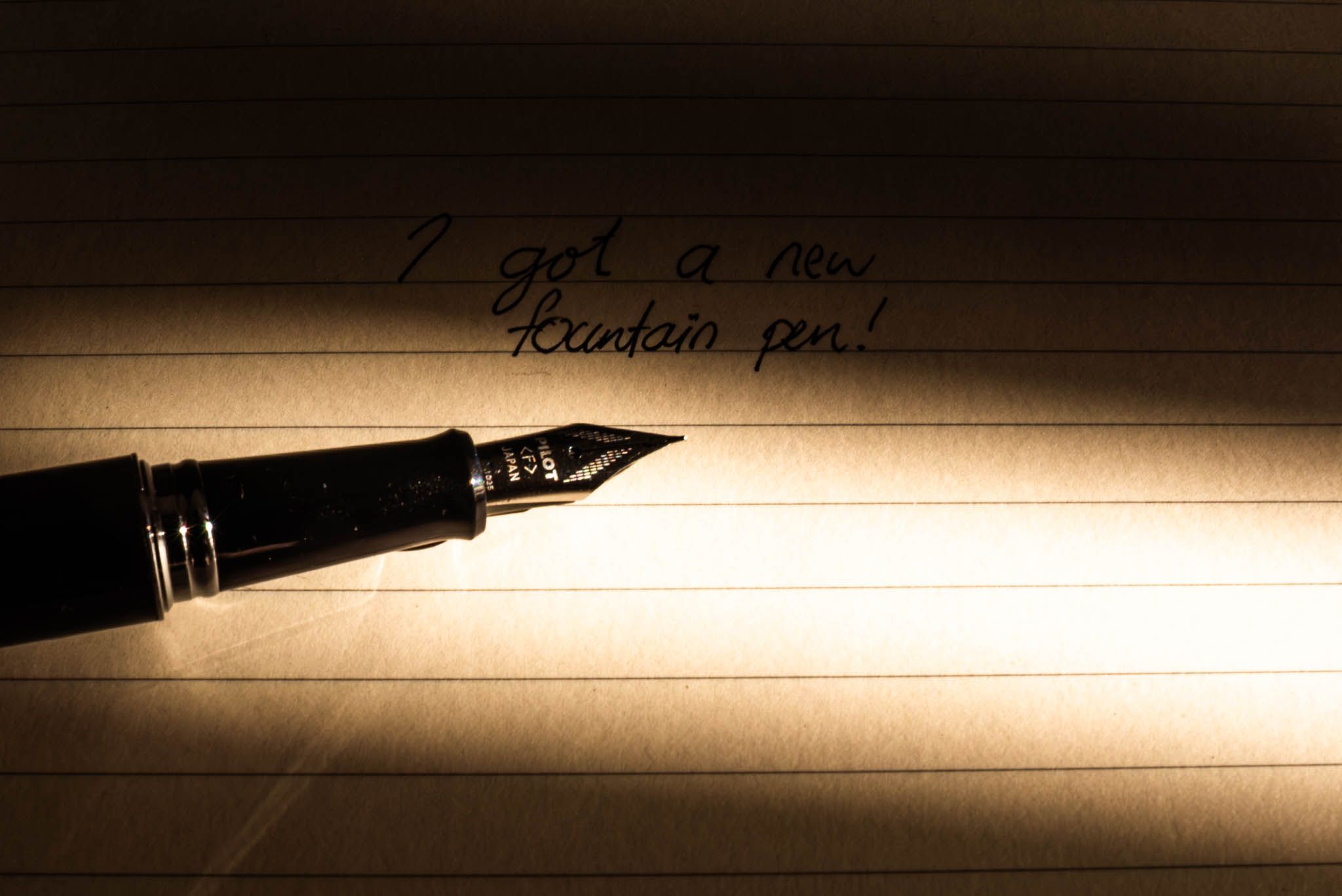 A fountain pen illuminated by natural light; in the background is lined paper with handwritten text saying, 'I got a new fountain pen'