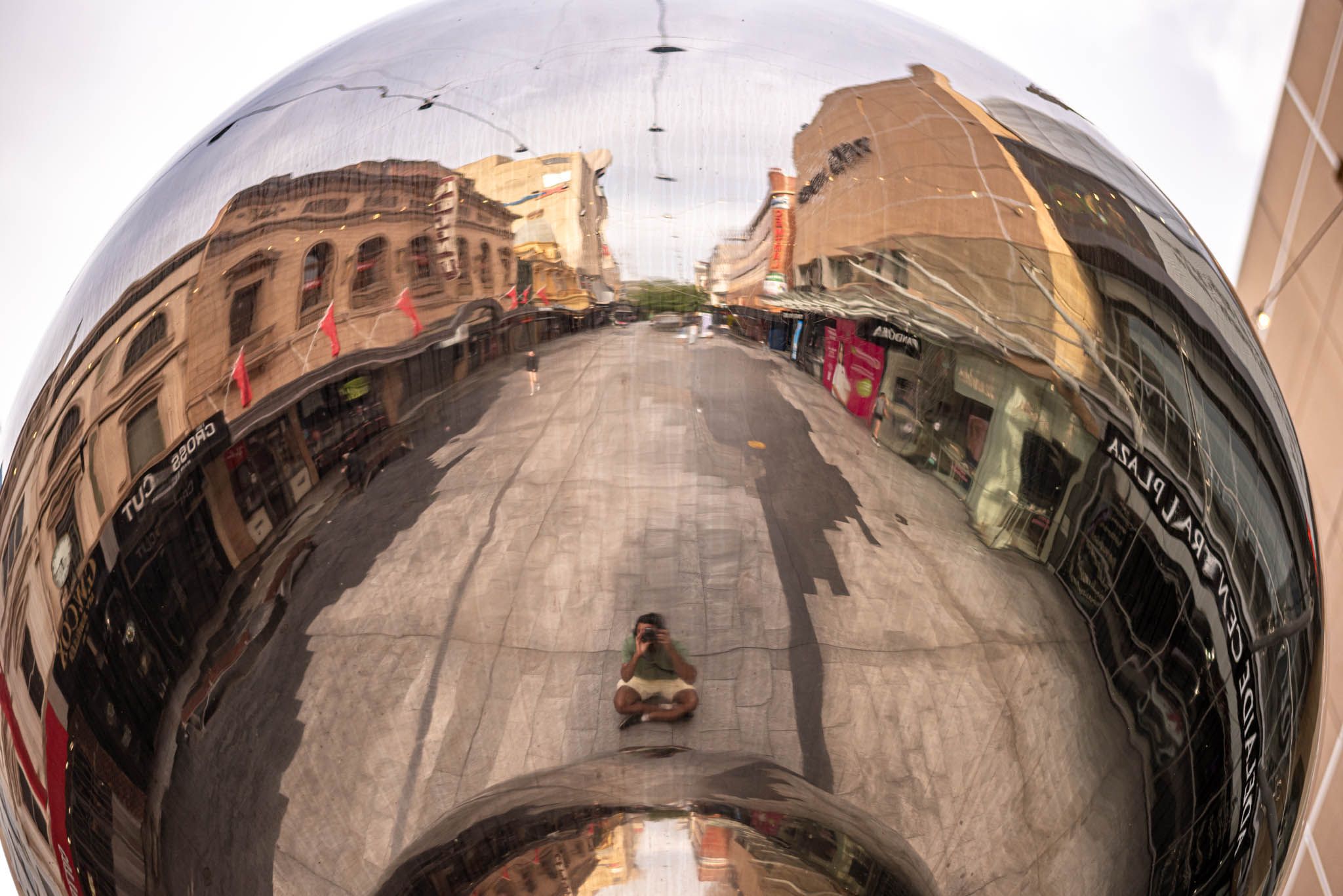 A large mirror ball with a individual wearing a green shirt taking a photo in the reflection